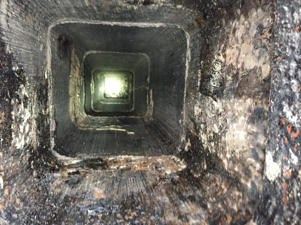 View looking down the inside of a sooted clay tile flue during a chimney inspection, with daylight visible at the damper